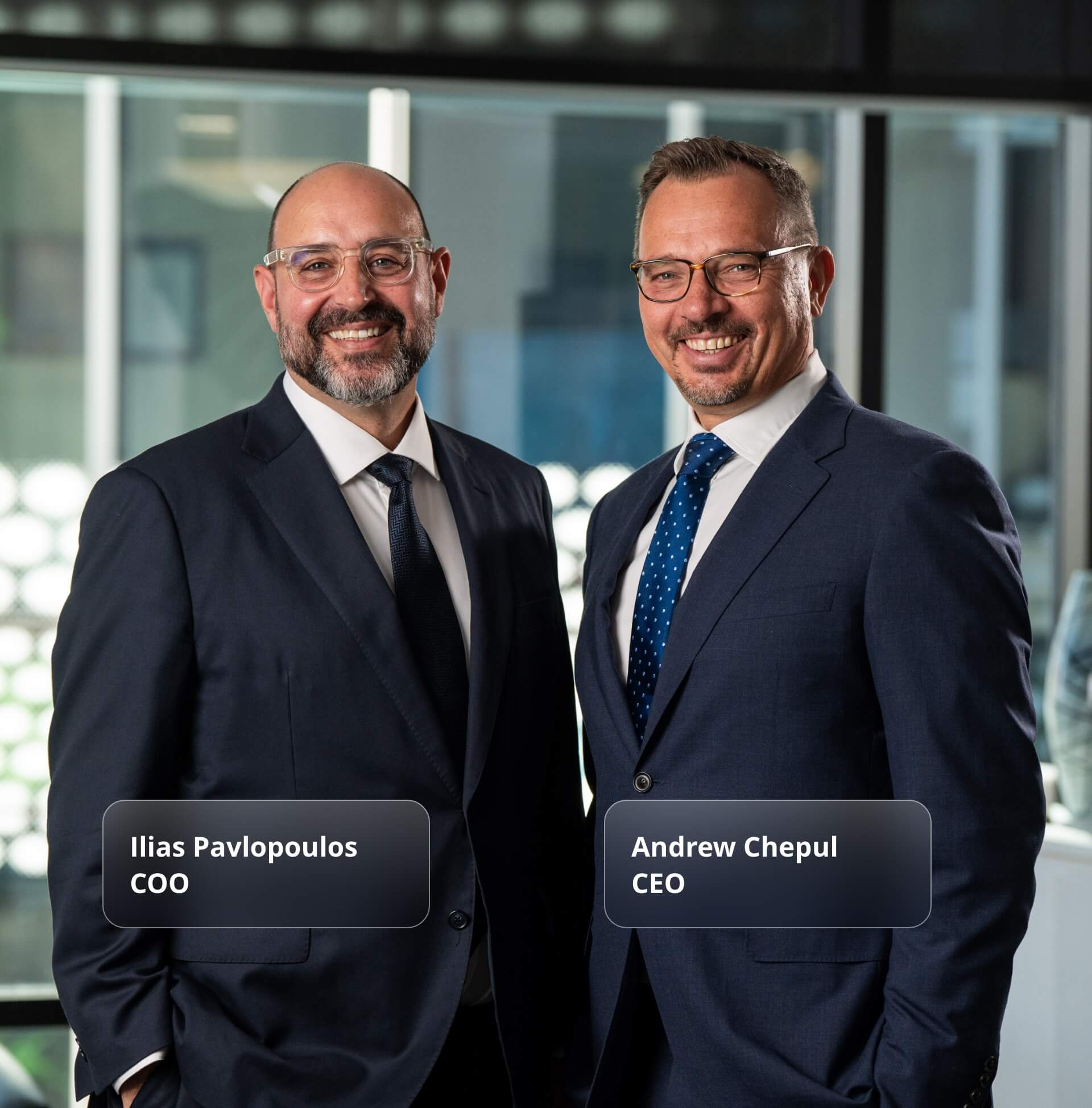 ColCap Cofounders in navy suits standing side by side in a modern office setting with glass walls and natural lighting, both wearing glasses and smiling confidently at the camera.