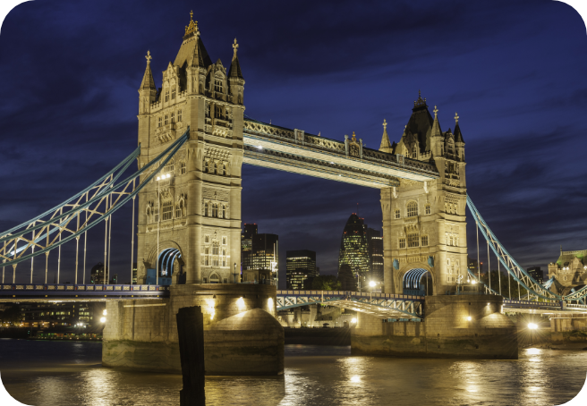 Illuminated Tower Bridge spanning the River Thames at dusk with the London city skyline visible through the bridge's iconic twin towers, reflecting golden lights on the water below against a deep blue evening sky.